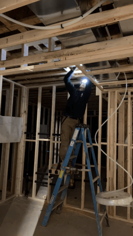 Construction worker on a blue ladder installs ceiling components in a wooden framed room.