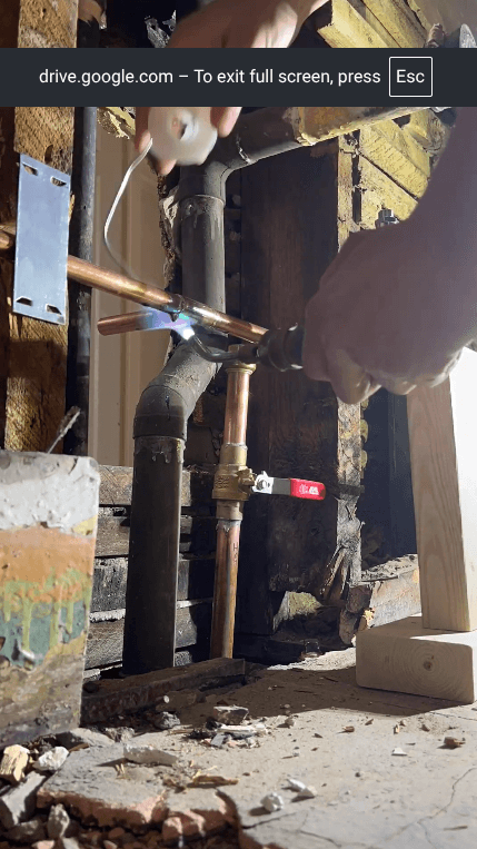 Person soldering a copper pipe joint with a blowtorch inside an exposed wooden wall.