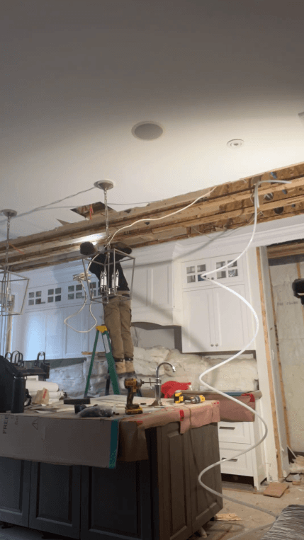 Worker on a ladder installs pendant lighting in a kitchen renovation with exposed ceiling beams.